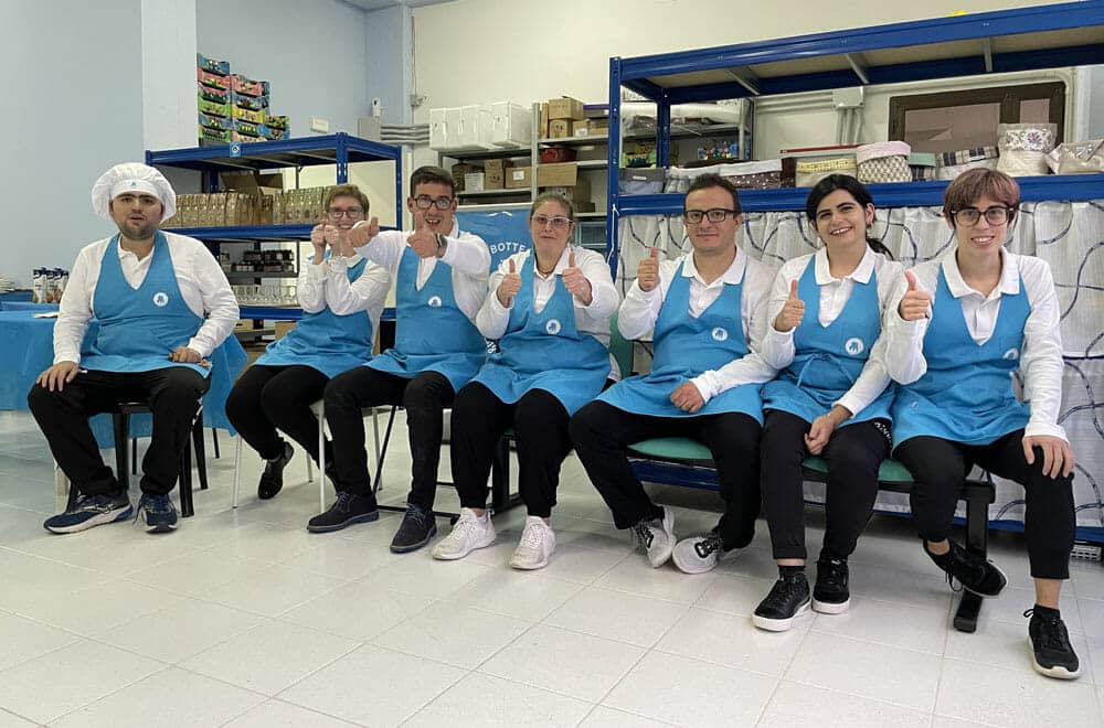 Un gruppo di pasticceri con uniformi bianche e grembiuli azzurri lavora insieme in un laboratorio di pasticceria, disponendo biscotti su teglie, con enfasi sulla collaborazione e il lavoro di squadra