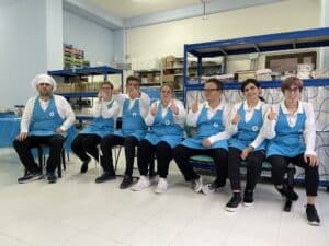 Un team di pasticceri in uniforme, con cappelli bianchi e grembiuli azzurri, siede in fila dando il pollice in su, dimostrando spirito di squadra e felicità nel loro ambiente di lavoro