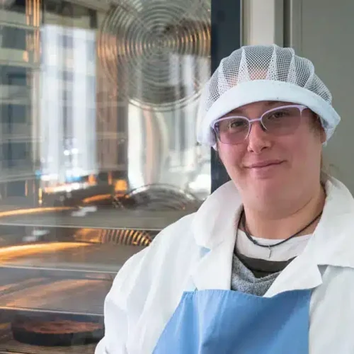 Una pasticcera con occhiali e un cappellino da chef sorride alla camera, davanti a un forno industriale con dolci in cottura, evidenziando un ambiente di pasticceria professionale