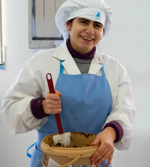 Letizia Una pasticcera sorridente in divisa da lavoro, con berretto e grembiule azzurro, mescola impasto al cioccolato in una ciotola in un laboratorio di pasticceria ben illuminato. L'ambiente riflette un'atmosfera accogliente e professionale